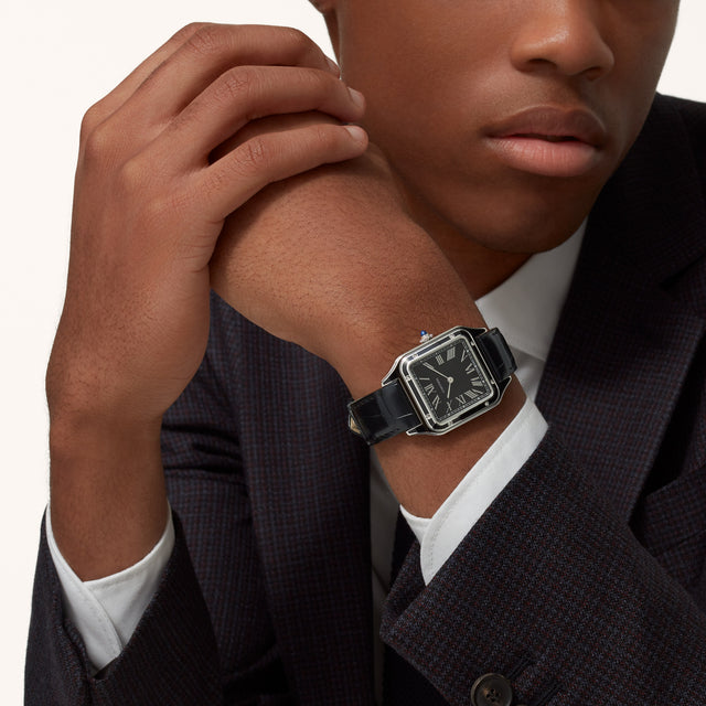 A person in a dark suit and white shirt rests their chin on their hand, highlighting the Cartier Santos-Dumont watch with its rectangular steel case and black leather strap.