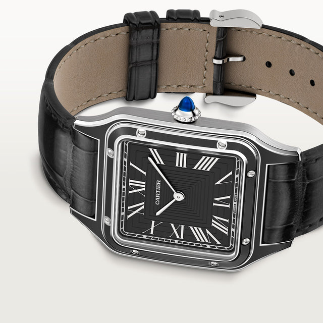 The Cartier Santos-Dumont watch features a black square face with Roman numerals, a steel case crowned with a blue gemstone, a black leather strap, and tan leather detailing on the watchback interior.