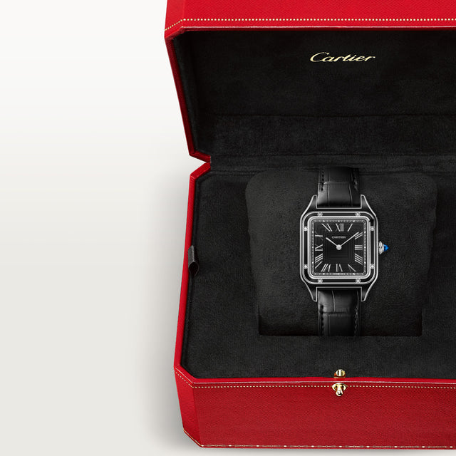 A Cartier Santos-Dumont watch with a black rectangular face, Roman numerals, and a sleek steel case is displayed in an open red Cartier box lined with velvet.