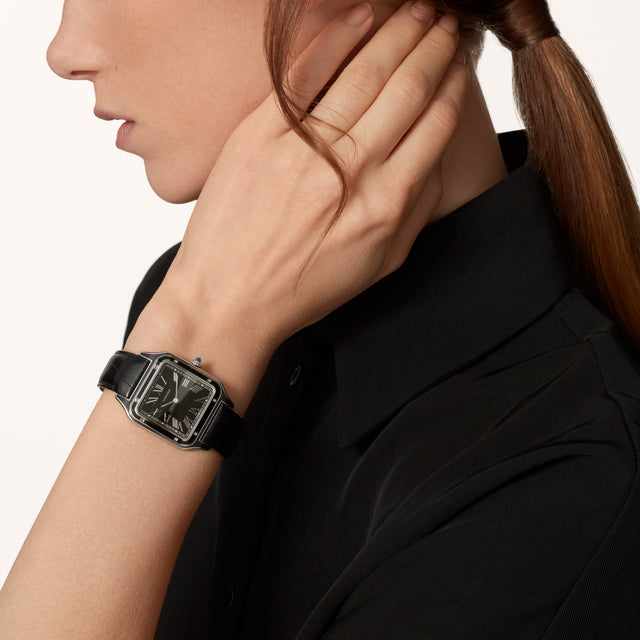 A woman in a black collared shirt touches her neck, showcasing the Cartier Santos-Dumont watch with a steel case and square black Roman numeral dial. Her hair is neatly tied back in a ponytail.