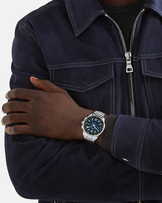 A person in a dark blue suede jacket with white stitching, arms crossed, wears a Jaeger-LeCoultre Polaris Chronograph with a striking blue lacquered dial, prominently displayed on their left wrist.